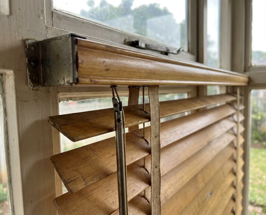 Timber venetian headrail up close on an older Northcliff window, tilt cords and ladder tapes at the bracket where most repairs begin