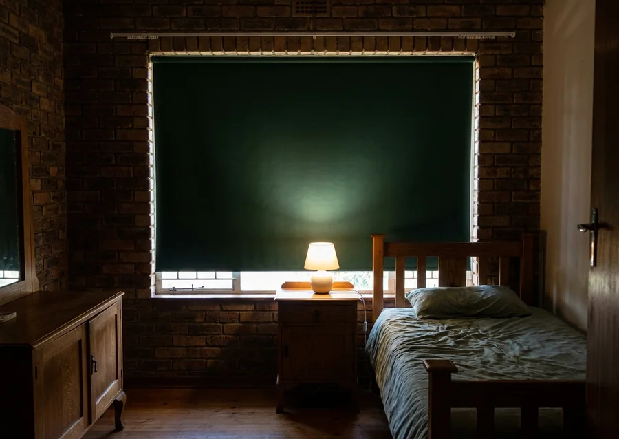 Forest-green blockout roller drawn over the steel-framed window of a face-brick Northcliff bedroom, afternoon daylight held to a thin glow at the edges