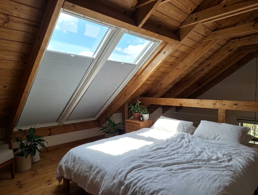 Two pleated skylight blinds half-drawn in their side tracks across sloped roof windows above a bed in a timber-ceilinged Northcliff loft